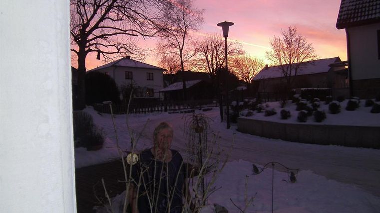 Eine verschneite Straße mit Häusern und Bäumen im Hintergrund. Der Himmel ist in sanften Rosa- und Violetttönen gefärbt.