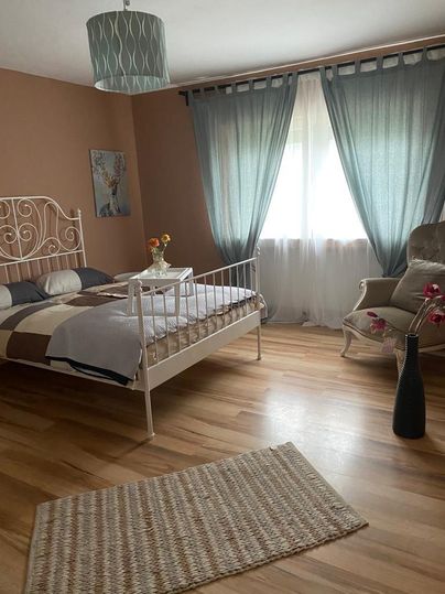 Ein gemütliches Schlafzimmer mit einem weißen Bett und einer bequemen Sessel. Die Fenster sind mit hellen Vorhängen dekoriert, und der Boden ist aus Holz.