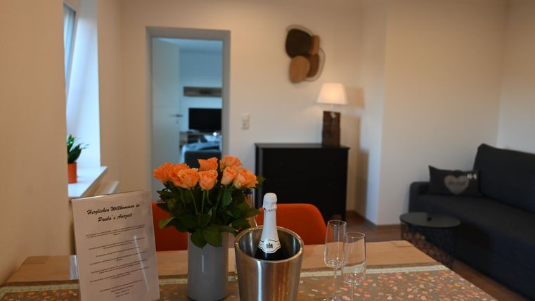 Ein gemütlicher Wohnbereich mit einem Tisch, auf dem eine Vase mit orangefarbenen Rosen steht. Im Hintergrund sind ein Sofa und ein Fernsehmöbel zu sehen.