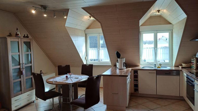 Eine moderne Küche mit schrägen Wänden und großen Fenstern. Es gibt einen Tisch mit Stühlen und eine Küchenzeile mit Geräten.
