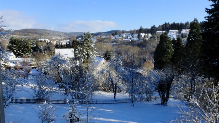 Eine winterliche Landschaft mit verschneiten Bäumen und Hügeln. Im Hintergrund sind Häuser und ein klarer blauer Himmel sichtbar.