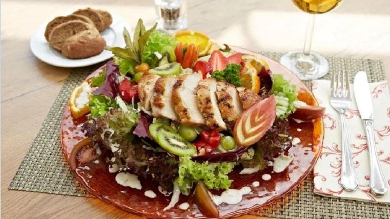 Ein bunter Salat mit gegrilltem Hähnchenfilet, frischem Gemüse und einer Vielzahl von Farben. Im Hintergrund sind ein Glas Getränk und ein Teller mit einer kleinen Portion Brot zu sehen.