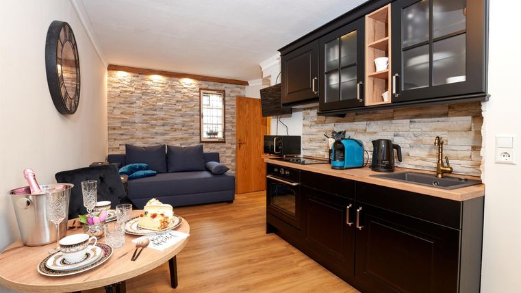 Eine moderne Küche mit schwarzen Schränken und Holzregal ist zu sehen. Daneben steht ein gemütliches Sofa und ein Tisch mit Geschirr.
