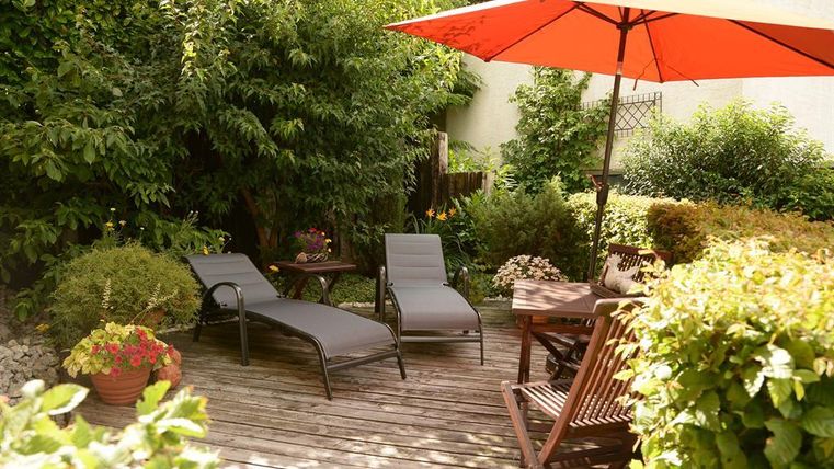 Ein einladender Garten mit zwei Liegen und einem roten Sonnenschirm. Umgeben von üppigem Grün und Blumen, lädt der Raum zum Entspannen ein.