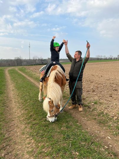 Ein Kind reitet auf einem Pferd und hebt die Arme in Jubel. Ein Erwachsener hält das Pferd an der Leine und lächelt.