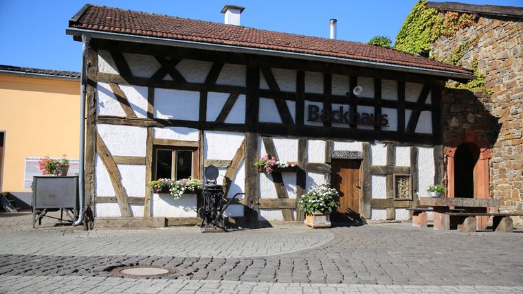 Fachwerkhaus mit der Aufschrift 'Backhaus', umgeben von Blumen und einem gepflasterten Platz.