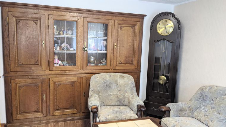 Ein gemütliches Wohnzimmer mit einem Holzschrank und zwei Stühlen. Im Hintergrund steht eine große Wanduhr.