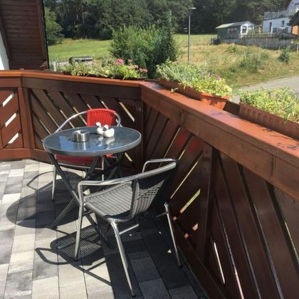 Ein gemütlicher Balkon mit einem runden Tisch und zwei Stühlen. Im Hintergrund sind grüne Wiesen und Bäume zu sehen.