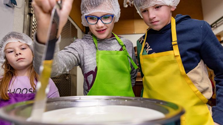Drei Kinder tragen Kochschürzen und Haarnetze, während sie gemeinsam an einem Topf arbeiten. Sie scheinen mit Interesse und Freude beim Kochen beschäftigt zu sein.
