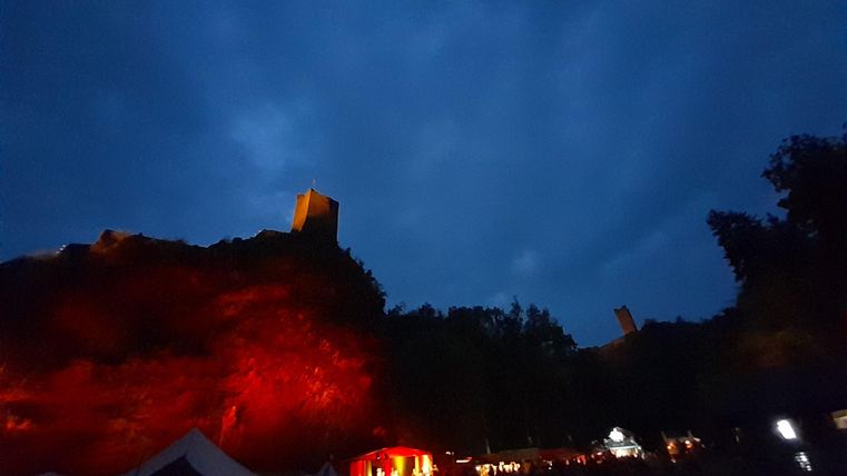Een indrukwekkend kasteel in de schemering, omringd door een donkere lucht en zacht licht. Het gebied eronder is verlicht en toont een feestelijke sfeer.