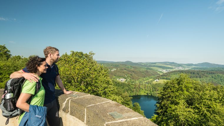 Zwei Personen stehen auf einem Aussichtspunkt und blicken auf eine grüne Landschaft mit einem See.