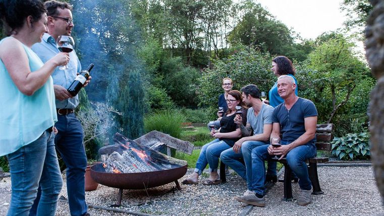 Eine gesellige Gruppe von Freunden versammelt sich um ein Feuer im Freien. Sie genießen Wein und die entspannte Atmosphäre in einer grünen Umgebung.