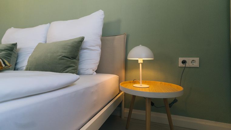 Ein modernes Schlafzimmer mit einem weißen Bett und gemütlichen Kissen. Auf dem Nachttisch steht eine stilvolle Lampe in warmem Licht.