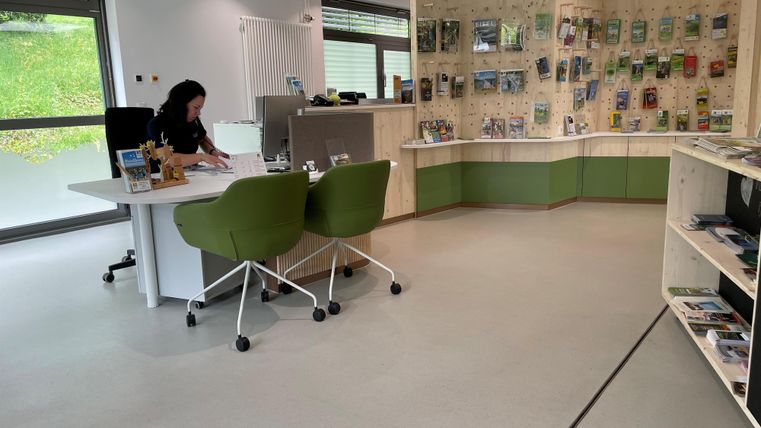 Ein modernes Büro mit einem Schreibtisch und zwei grünen Stühlen. An der Wand sind Informationsmaterialien und Broschüren ausgestellt.