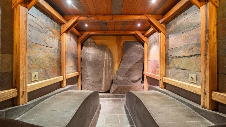 Ein entspannender Wellnessbereich mit zwei Ruheliegen aus Stein. Die Wände bestehen aus natürlichem Gestein und Holz, was eine gemütliche Atmosphäre schafft.