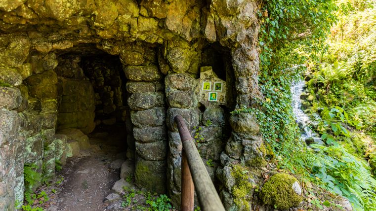 A rocky entrance, carved into the cliff face, leading to a small cave gallery with a path secured by a handrail, surrounded by moss and lush greenery; to the right, a waterfall cascades down between the rocks.