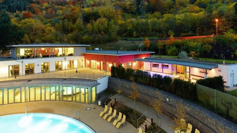 Een modern wellnesshotel met een uitnodigende buitenzwembad. De omgeving wordt gekenmerkt door groene heuvels en kleurrijk loof.