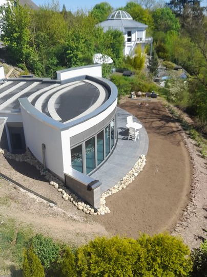 Een modern huis met een gebogen gevel en grote ramen. Omgeven door een groen landschap en een aangelegde tuin.