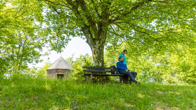 Zwei Personen sitzen lächelnd auf einer Bank im grünen unter einem Baum. Im Hintergrund befindet sich eine kleine Steinhütte.