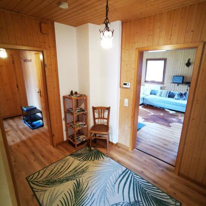 Ein einladender Flur mit Holzverkleidung, einer Lampe und einem Stuhl. Auf dem Boden liegt ein Teppich mit Palmenmotiv.