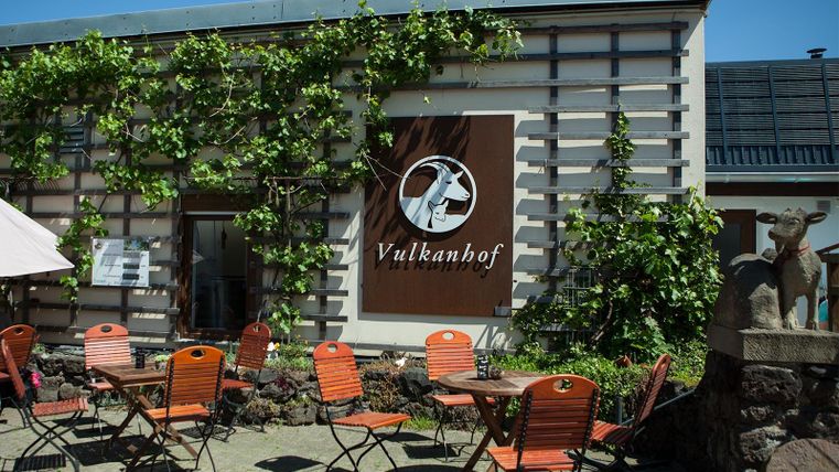 Ein rustikales Gebäude mit dem Schild "Vulkanhof" ist von Pflanzen umgeben. Vor dem Gebäude stehen orangefarbene Stühle und Tische.