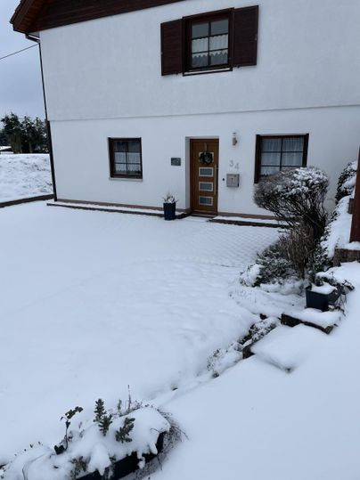 Ein Haus im Schnee mit einer verschneiten Auffahrt. Der Garten ist mit Schnee bedeckt und es stehen einige Pflanzen in Töpfen.