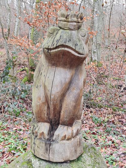 Eine Holzskulptur eines Frosches steht in einem Wald. Die Umgebung ist voller Bäume und Laub.