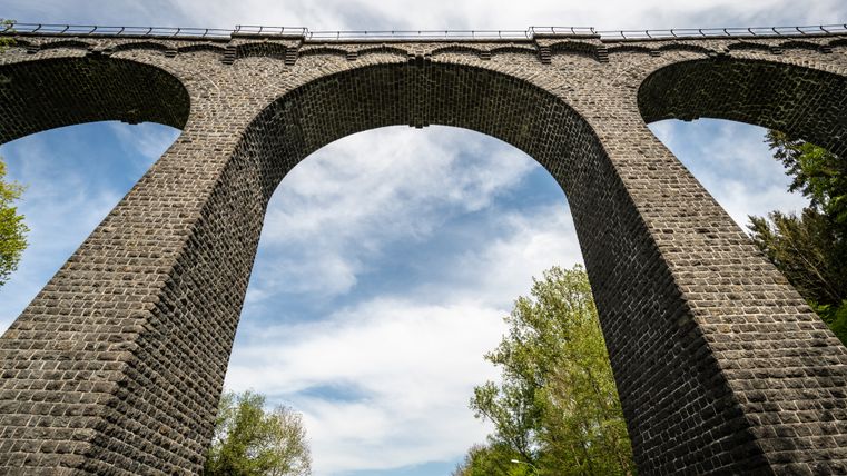 Ansicht eines Viadukts von unten mit blauem Himmel und Bäumen im Hintergrund.