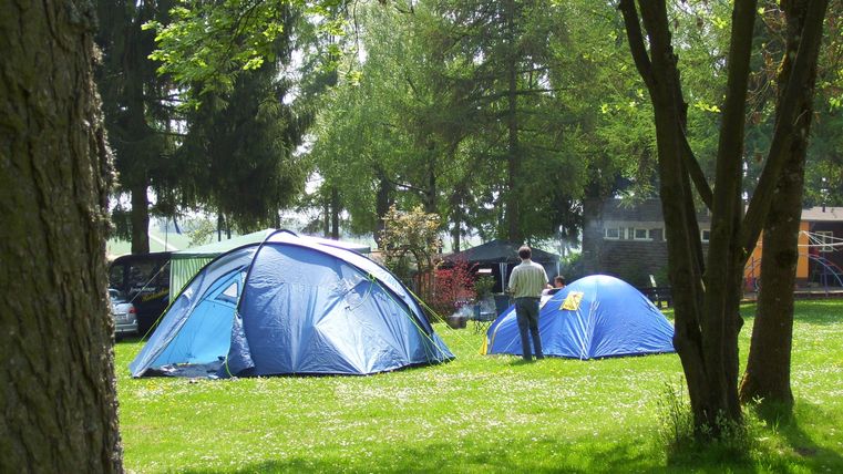 Ein Campingplatz mit blauen Zelten auf grünem Gras. Umgeben von Bäumen und einem sonnigen Himmel.