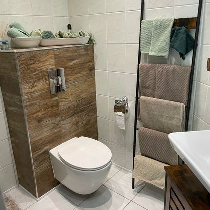 Ein modernes Badezimmer mit einer weißen Toilette und einer Holzablage. Handtücher in sanften Farben hängen an einem Ständer neben einem Waschbecken.