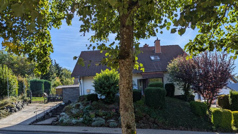 Ein kleines Haus umgeben von Bäumen und gepflegtem Garten. Der Himmel ist klar und sonnig.