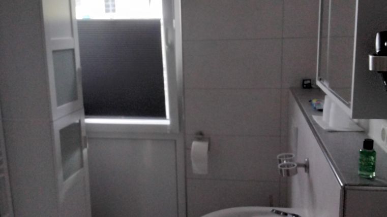 Ein modernes Badezimmer mit einem Waschbecken, einer Dusche und einem Fenster. Die Wände sind hell gefliest und der Boden ist grau.