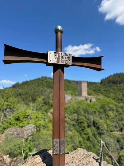 Ein Wegweiser auf einem Hügel mit Blick auf eine grüne Landschaft und eine Burgruine im Hintergrund. Der Himmel ist blau mit wenigen Wolken.