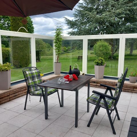 Ein gemütlicher Essbereich im Freien mit einem Tisch und zwei Stühlen. Im Hintergrund sind grüne Pflanzen und eine ruhige Landschaft zu sehen.