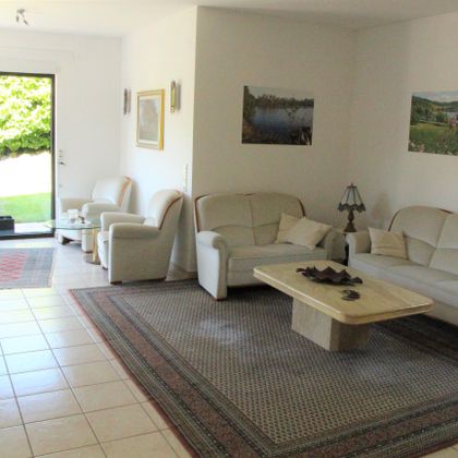Ein helles Wohnzimmer mit weißen Sofas und einem Tisch in der Mitte. Große Fenster bieten Blick auf den Garten.