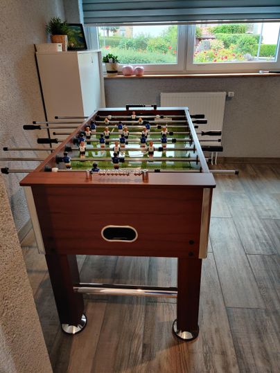 Ein moderner Tischkicker aus Holz steht in einem Raum mit Fenster. Die Umgebung ist hell und freundlich, ideal für Spiele und Freizeitaktivitäten.