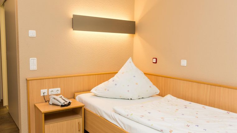 Ein einfaches Schlafzimmer mit einem Holzbett und einer Kissenform im Dreieck. Die Wände sind in einem neutralen Farbton gestrichen, und die Beleuchtung ist sanft.