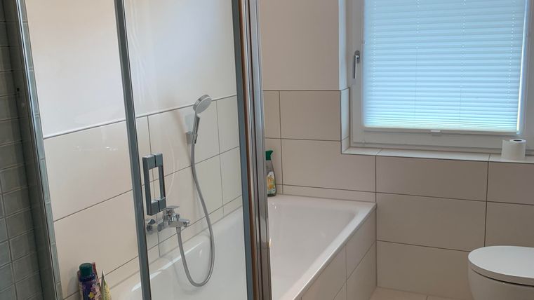 Ein modernes Badezimmer mit einer Dusche und einer Badewanne. Es gibt große Fliesen und ein Fenster, das natürliches Licht hereinscheinen lässt.