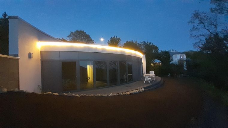 Ein modernes Haus mit sanften Rundungen leuchtet sanft in der Dämmerung. Es hat große Fenster und einen gepflegten Außenbereich.