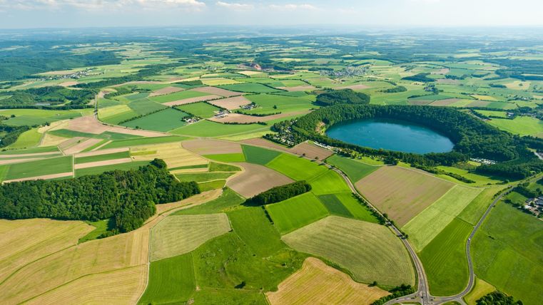 Eine weite Landschaft mit grünen Feldern und Wäldern. Im Mittelpunkt befindet sich ein runder See, umgeben von landwirtschaftlichen Flächen.