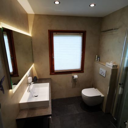 Ein modernes Badezimmer mit einem Waschbecken, einer Toilette und einer Dusche. Das Fenster lässt Tageslicht herein und sorgt für eine helle Atmosphäre.