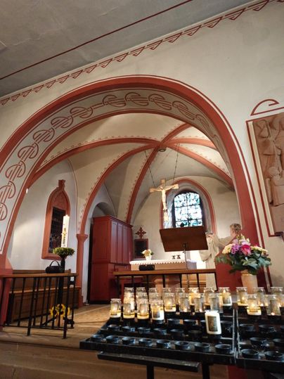 Eine ruhige Kapelle mit kunstvoll gestalteten Wänden und einem Altar. Im Vordergrund stehen viele brennende Kerzen und frische Blumen.