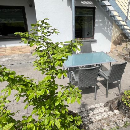 Ein gemütlicher Außenbereich mit einem Glastisch und vier Stühlen. Im Hintergrund sieht man ein helles Haus und grünes Laub.