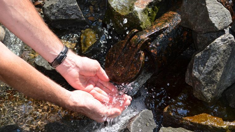 Eine Hand, die Wasser aus einem kleinen Bach auffängt, umgeben von steinigen Ufern. Das Wasser spritzt klar und sprudelnd zwischen den Steinen.