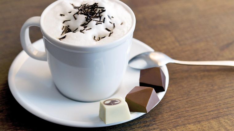 Eine Tasse Kaffee mit Milchschaum und Schokoladenraspeln. Dazu liegen drei verschiedene Pralinen auf einem Unterteller.