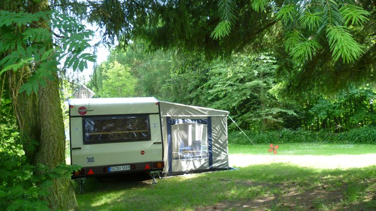 Ein Campinganhänger auf einer Wiese, umgeben von Bäumen. Das Gelände ist grün und friedlich.