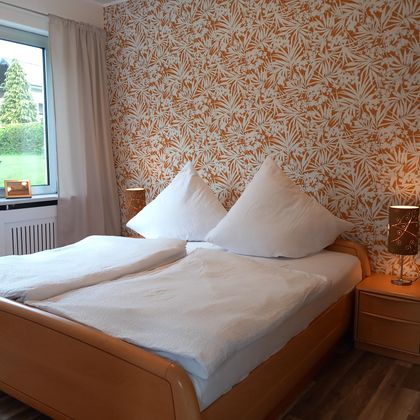 A cozy bedroom with a double bed and large pillows. The walls are decorated with a floral pattern and a window lets in daylight.