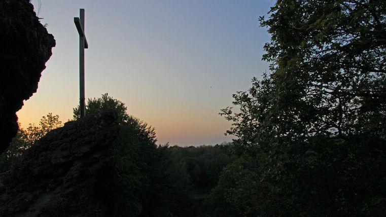 Ein Holzkreuz steht auf einem Felsen, umgeben von Bäumen. Der Himmel färbt sich bei Sonnenuntergang in sanften Tönen.