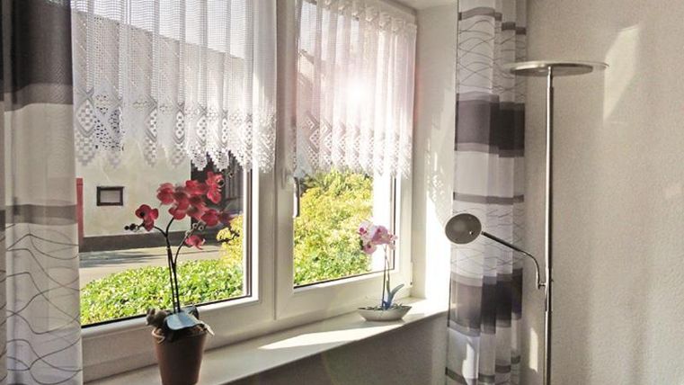 Ein heller Raum mit zwei Fenstern, die mit Gardinen dekoriert sind. Auf der Fensterbank stehen Pflanzen und ein Stehlampe.