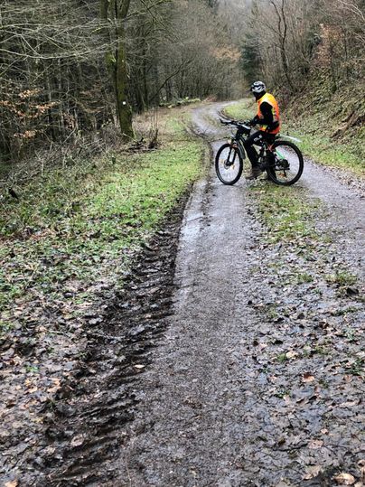 Ein Mountainbiker steht auf einem matschigen Waldweg. Die Umgebung ist von Bäumen umgeben und zeigt eine ländliche Atmosphäre.